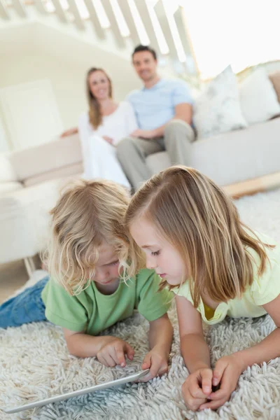 Rodzeństwo, za pomocą tabletki na dywanie z rodzicami za nimi — Zdjęcie stockowe