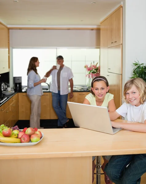 Rodzeństwo z laptopa w kuchni z rodzicami za nimi — Zdjęcie stockowe