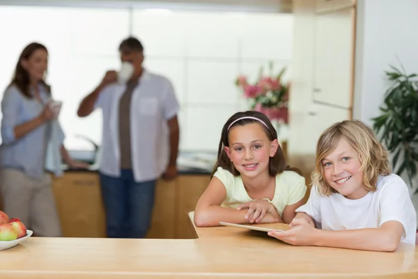Братья и сестры с помощью планшета на кухне с родителями позади них — стоковое фото
