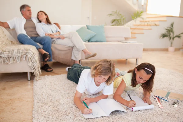 Братья и сёстры делают домашнее задание на полу с родителями позади них — стоковое фото