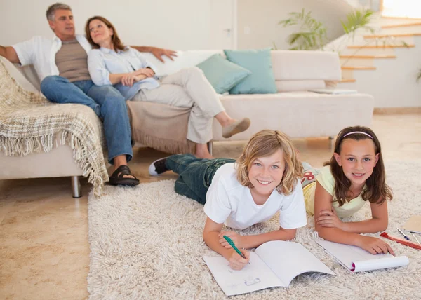 Frères et sœurs faisant leurs devoirs sur le tapis avec leurs parents derrière eux — Photo