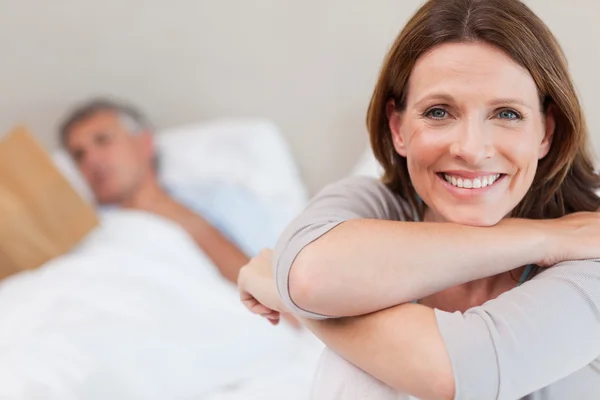 Улыбающаяся женщина в постели с читающим мужем на заднем плане — стоковое фото