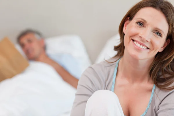 Érett nő ágyon olvasó férje háta mögött mosolygó — Stock Fotó