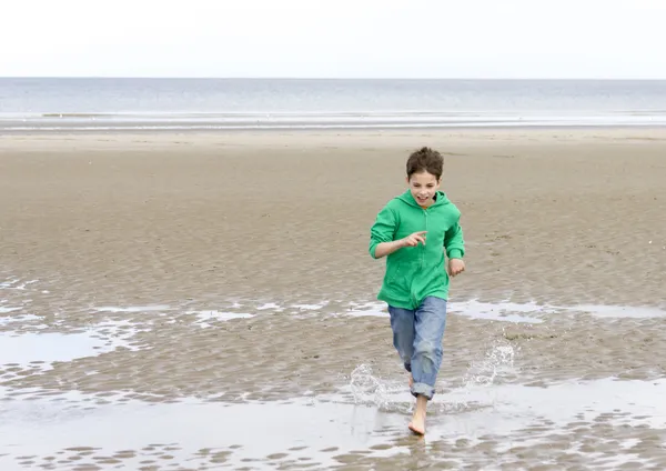 stock image The boy in a green hoodie, running barefoot on the beach at low