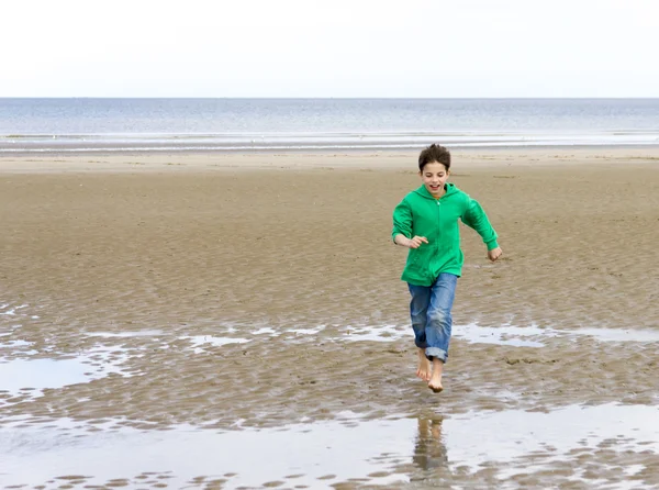 stock image The boy in a green hoodie, running barefoot on the beach