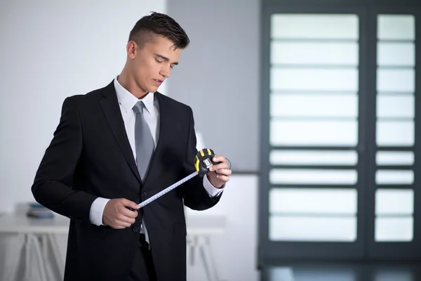 Joven hombre de negocios en un traje sosteniendo y mirando una cinta métrica —  Fotos de Stock