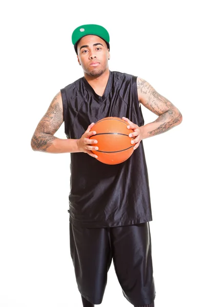 Retrato de estudio del jugador de baloncesto con gorra verde de pie y sosteniendo la pelota aislada en blanco. Tatuajes en sus brazos . —  Fotos de Stock