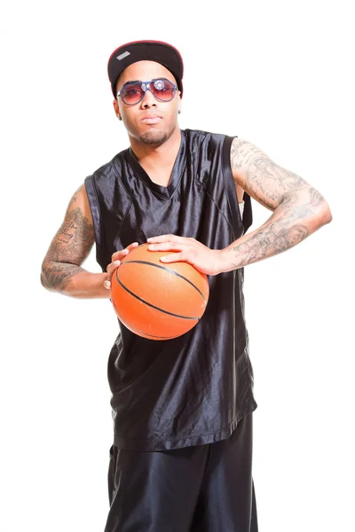 篮球运动员戴帽和太阳镜站和持有球上白色隔离室画像。他手臂上的纹身. — 图库照片