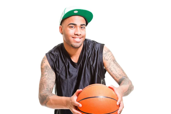 Retrato de estudio del jugador de baloncesto con gorra verde de pie y sosteniendo la pelota aislada en blanco. Tatuajes en sus brazos . — Foto de Stock
