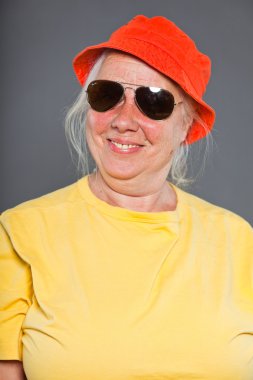 mutlu ve komik üst düzey kadın sarı tişört ve turuncu şapka ve güneş gözlüğü takıyor. serin ve kalça. izole üzerinde gri Studio vurdu.