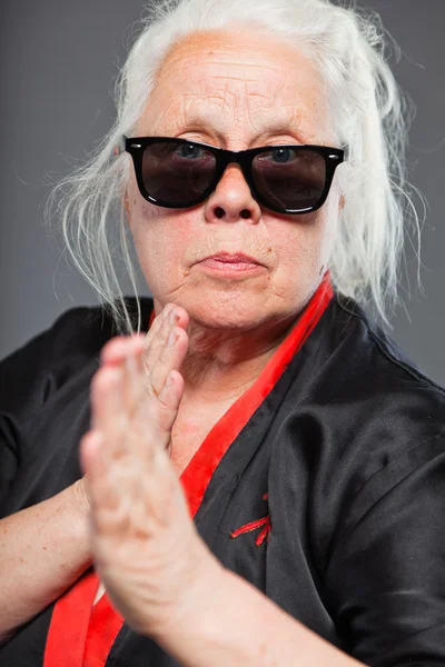 Starszy kobieta z długie siwe włosy, noszenie kimono czarne i czerwone i czarne okulary. robi karate porusza. Studio strzał na białym tle na szarym tle. — Zdjęcie stockowe