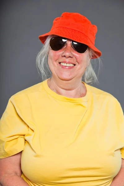 Femme âgée drôle et heureuse portant chemise jaune et chapeau orange et lunettes de soleil. Cool et branché. Studio tourné isolé sur gris . — Photo
