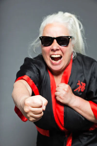 Mujer mayor con el pelo largo y gris usando kimono negro y rojo y gafas de sol negras. Haciendo movimientos de karate. Estudio plano aislado sobre fondo gris . — Foto de Stock