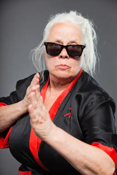 Äldre kvinna med långt grått hår, klädd i svart och röd kimono och svarta solglasögon. gör karate flyttar. Studio skott isolerade på grå bakgrund. — Stockfoto