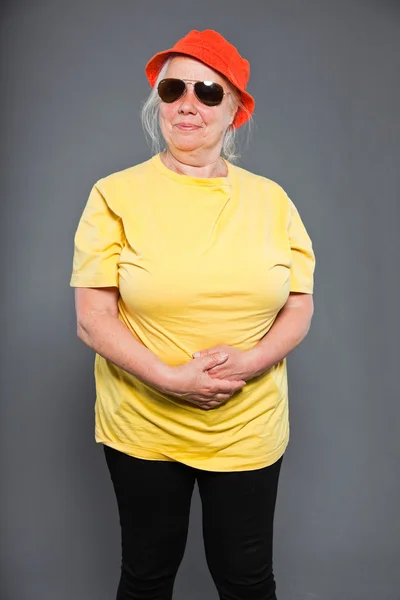Mutlu ve komik üst düzey kadın sarı tişört ve turuncu şapka ve güneş gözlüğü takıyor. serin ve kalça. izole üzerinde gri Studio vurdu. — Stok fotoğraf