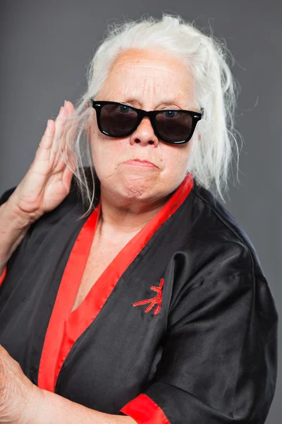Femme âgée aux longs cheveux gris portant du kimono noir et rouge et des lunettes de soleil noires. Faire des mouvements de karaté. Studio tourné isolé sur fond gris . — Photo