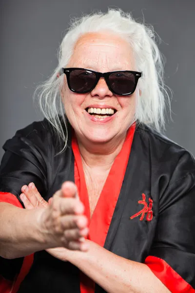 Äldre kvinna med långt grått hår, klädd i svart och röd kimono och svarta solglasögon. gör karate flyttar. Studio skott isolerade på grå bakgrund. — Stockfoto