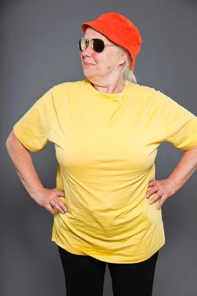 Lustige und fröhliche Seniorin mit gelbem Hemd und orangefarbenem Hut und Sonnenbrille. cool und hip. Studioaufnahme isoliert auf grau. — Stockfoto