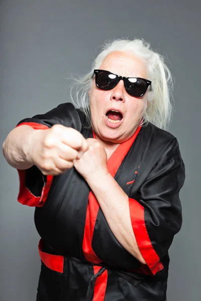 Senior woman with long grey hair wearing black and red kimono and black sunglasses. Doing karate moves. Studio shot isolated on grey background. — Stock Photo, Image