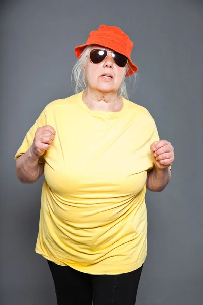 Lustige und fröhliche Seniorin mit gelbem Hemd und orangefarbenem Hut und Sonnenbrille. cool und hip. Studioaufnahme isoliert auf grau. — Stockfoto