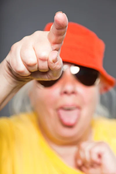 Αστεία και ευτυχισμένη γυναίκα ανώτερος φοράει κίτρινο πουκάμισο και το πορτοκαλί καπέλο και γυαλιά ηλίου. δροσερός και του ισχίου. στούντιο πυροβόλησε απομονωμένες σε γκρι. — Φωτογραφία Αρχείου