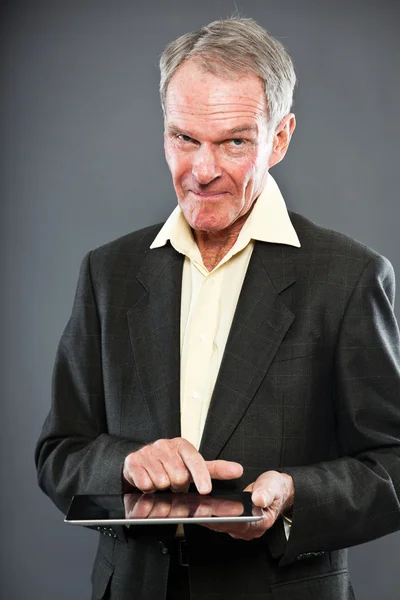 Expressieve goed uitziende senior man in donker pak tegen grijs muur. met behulp van Tablet PC. grappig en karakteristiek. goed gekleed. studio opname. — Stockfoto