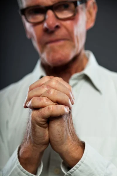 Expressieve goed uitziende senior man met bril tegen grijs muur. handen bidden. geestelijke en karakteristieke. goed gekleed. studio opname. — Stockfoto
