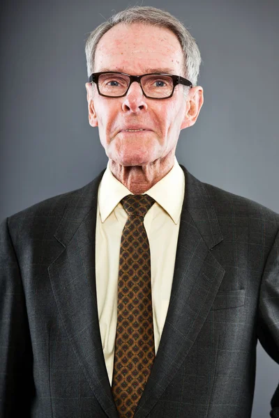 Ausdrucksstarker gut aussehender älterer Herr im dunklen Anzug gegen graue Wand. Brille auf. Witzig und charakteristisch. Gut angezogen. Studioaufnahme. — Stockfoto