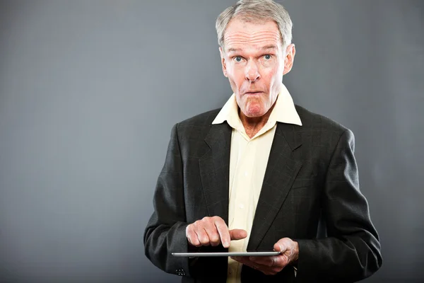 Expressieve goed uitziende senior man in donker pak tegen grijs muur. met behulp van Tablet PC. grappig en karakteristiek. goed gekleed. studio opname. — Stockfoto