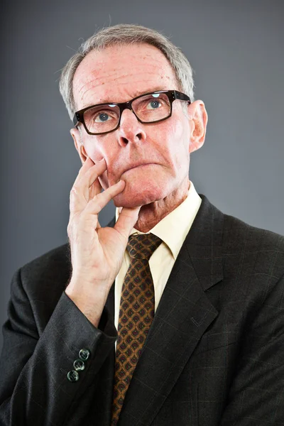 Expressive good looking senior man in dark suit against grey wall. Wearing glasses. Funny and characteristic. Well dressed. Studio shot. — Stock Photo, Image