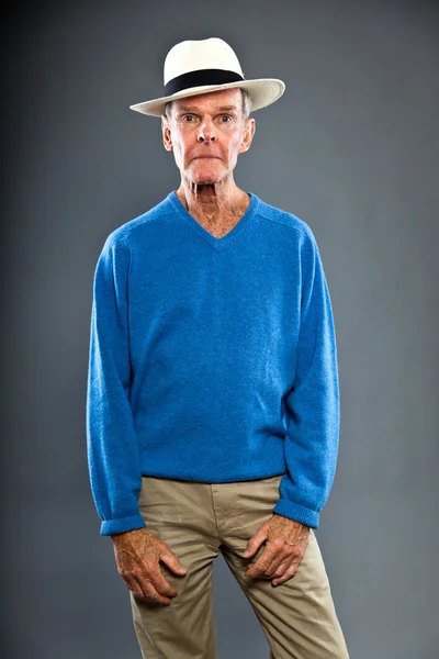 Expressive good looking senior man against grey wall. Wearing hat. Funny and characteristic. Well dressed. Blue sweater. Studio shot. — Stock Photo, Image