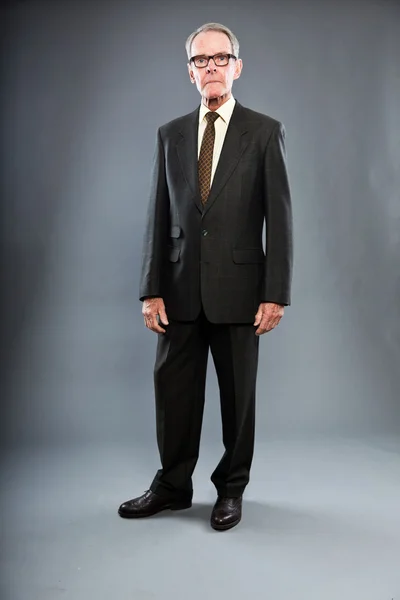 Uttrycksfulla snygg äldre man i mörk kostym mot grå vägg. glasögon. rolig och karakteristiska. välklädd. Studio skott. — Stockfoto