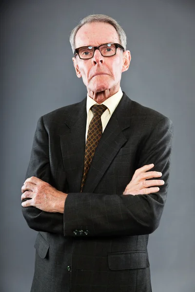Expressive good looking senior man in dark suit against grey wall. Wearing glasses. Funny and characteristic. Well dressed. Studio shot. — Stock Photo, Image