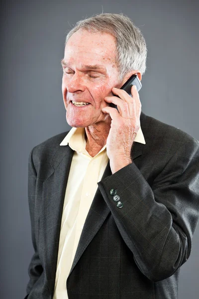 Ausdrucksstarker gut aussehender älterer Herr im dunklen Anzug gegen graue Wand. Telefonieren mit dem Handy. Witzig und charakteristisch. Gut angezogen. Studioaufnahme. — Stockfoto