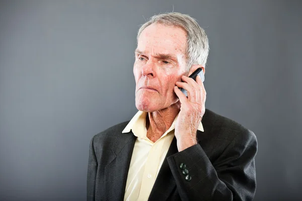Expressieve goed uitziende senior man in donker pak tegen grijs muur. bellen met mobiele telefoon. grappig en karakteristiek. goed gekleed. studio opname. — Stockfoto