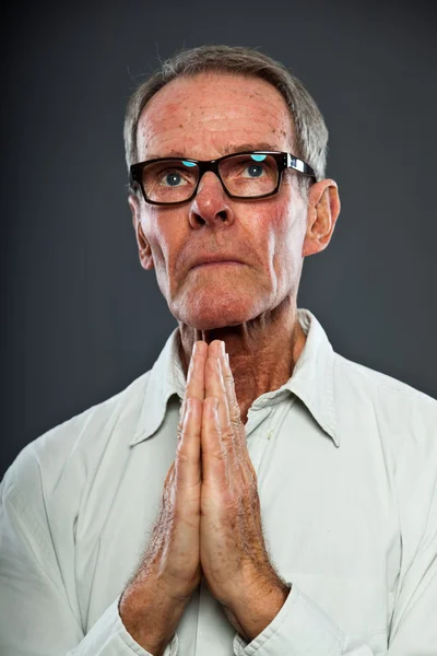 Ausdrucksstarker gut aussehender älterer Herr mit Brille vor grauer Wand. Hände beten. spirituell und charakteristisch. Gut angezogen. Studioaufnahme. — Stockfoto