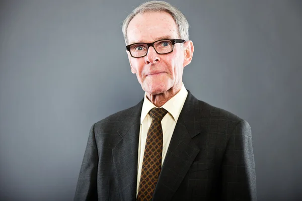 Expressive good looking senior man in dark suit against grey wall. Wearing glasses. Funny and characteristic. Well dressed. Studio shot. — Stock Photo, Image