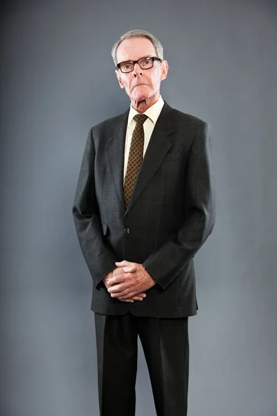 Ausdrucksstarker gut aussehender älterer Herr im dunklen Anzug gegen graue Wand. Brille auf. Witzig und charakteristisch. Gut angezogen. Studioaufnahme. — Stockfoto