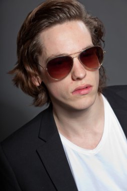 uzun kahverengi saçlı, beyaz gömlek ve mavi ceket ve gri arka plan üzerinde izole güneş gözlüğü takan yakışıklı genç adam. moda studio vurdu. etkileyici yüz.