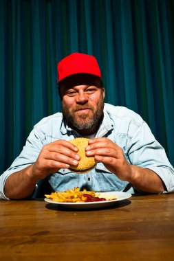 fast food yemek yeme sakallı adam. hamburger ve patates kızartması tadını çıkarıyor. kırmızı bere ile kamyon şoförü.