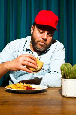 fast food yemek yeme sakallı adam. hamburger ve patates kızartması tadını çıkarıyor. kırmızı bere ile kamyon şoförü.