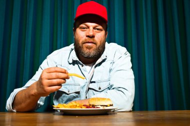 fast food yemek yeme sakallı adam. hamburger ve patates kızartması tadını çıkarıyor. kırmızı bere ile kamyon şoförü.