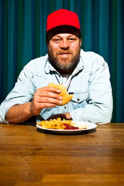 fast food yemek yeme sakallı adam. hamburger ve patates kızartması tadını çıkarıyor. kırmızı bere ile kamyon şoförü.