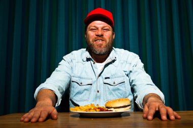 fast food yemek yeme sakallı adam. hamburger ve patates kızartması tadını çıkarıyor. kırmızı bere ile kamyon şoförü.