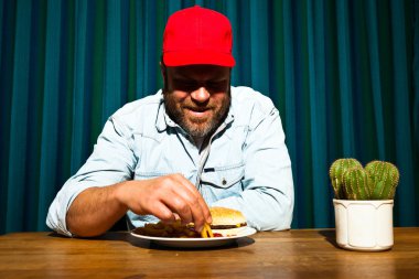 fast food yemek yeme sakallı adam. hamburger ve patates kızartması tadını çıkarıyor. kırmızı bere ile kamyon şoförü.