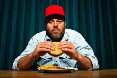 fast food yemek yeme sakallı adam. hamburger ve patates kızartması tadını çıkarıyor. kırmızı bere ile kamyon şoförü.