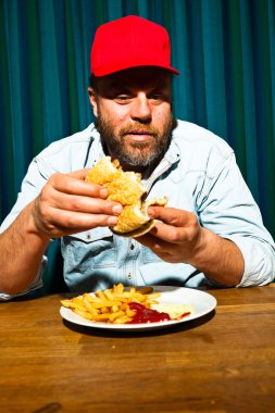 fast food yemek yeme sakallı adam. hamburger ve patates kızartması tadını çıkarıyor. kırmızı bere ile kamyon şoförü.