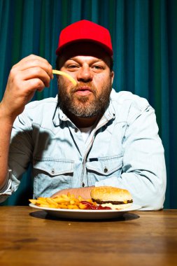 fast food yemek yeme sakallı adam. hamburger ve patates kızartması tadını çıkarıyor. kırmızı bere ile kamyon şoförü.