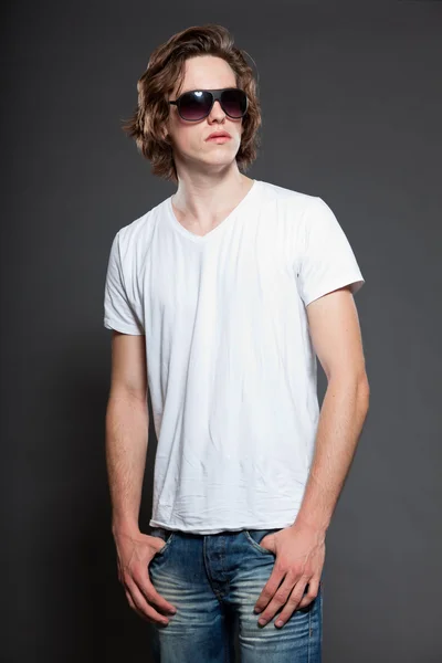 Beau jeune homme aux cheveux longs bruns et aux lunettes de soleil isolées sur fond gris. Studio de mode tourné . — Photo