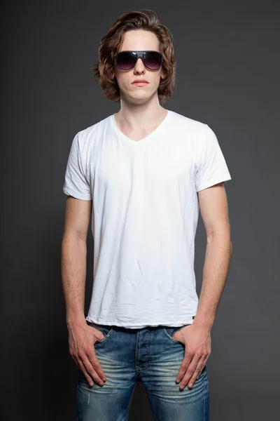 Beau jeune homme aux cheveux longs bruns et aux lunettes de soleil isolées sur fond gris. Studio de mode tourné . — Photo